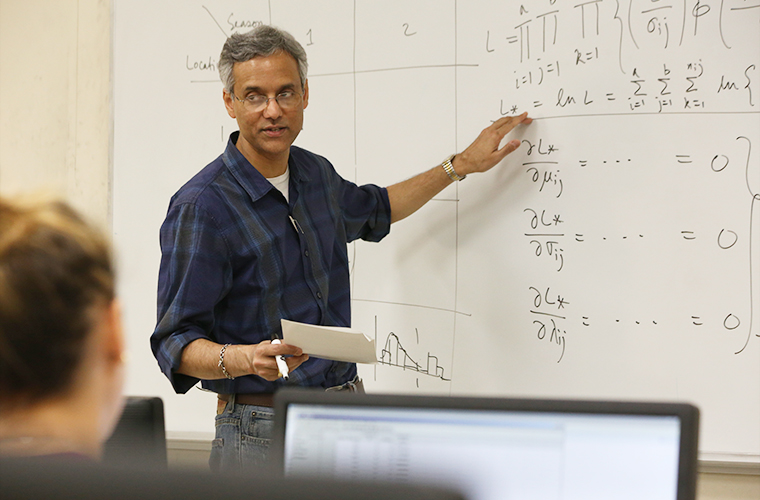 Math Professor faces students, pointing to math equations on a whiteboard.