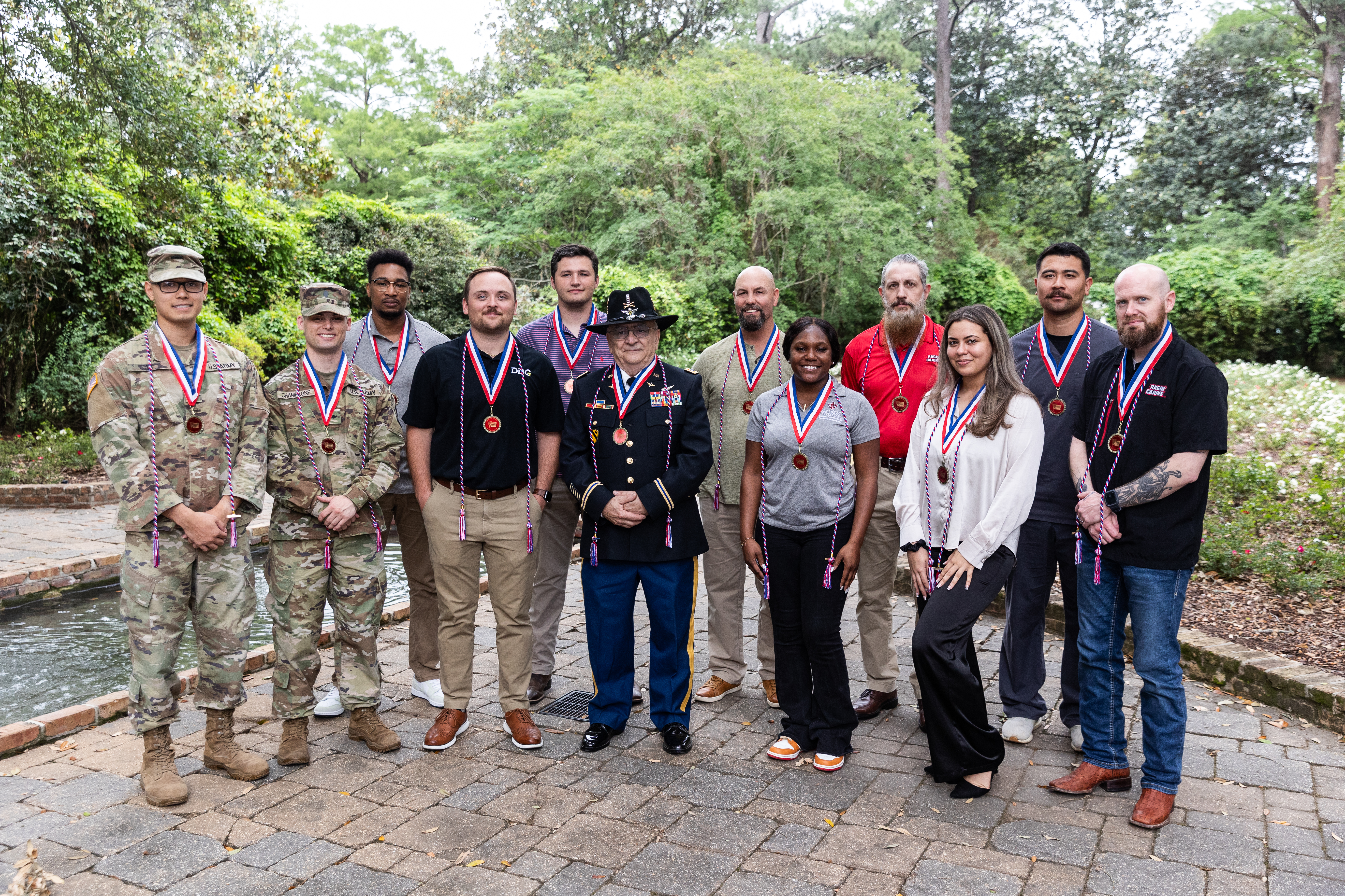 12 people stand on cobblestone pavement in front of green trees and a small pond, each with a red white and blue medallion and cord