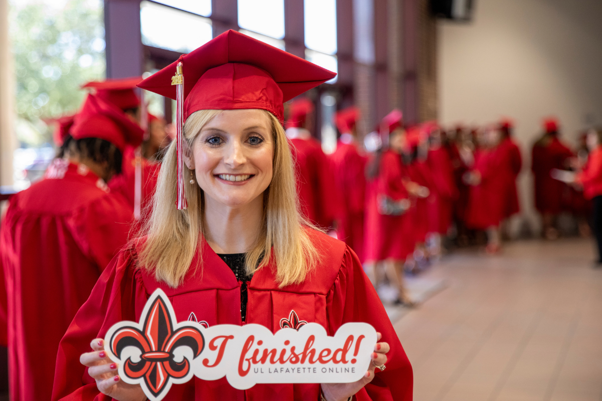 Online HPW graduate holds a UL Lafayette sign that reads 'I finished!' while smiling in her cap and gown.
