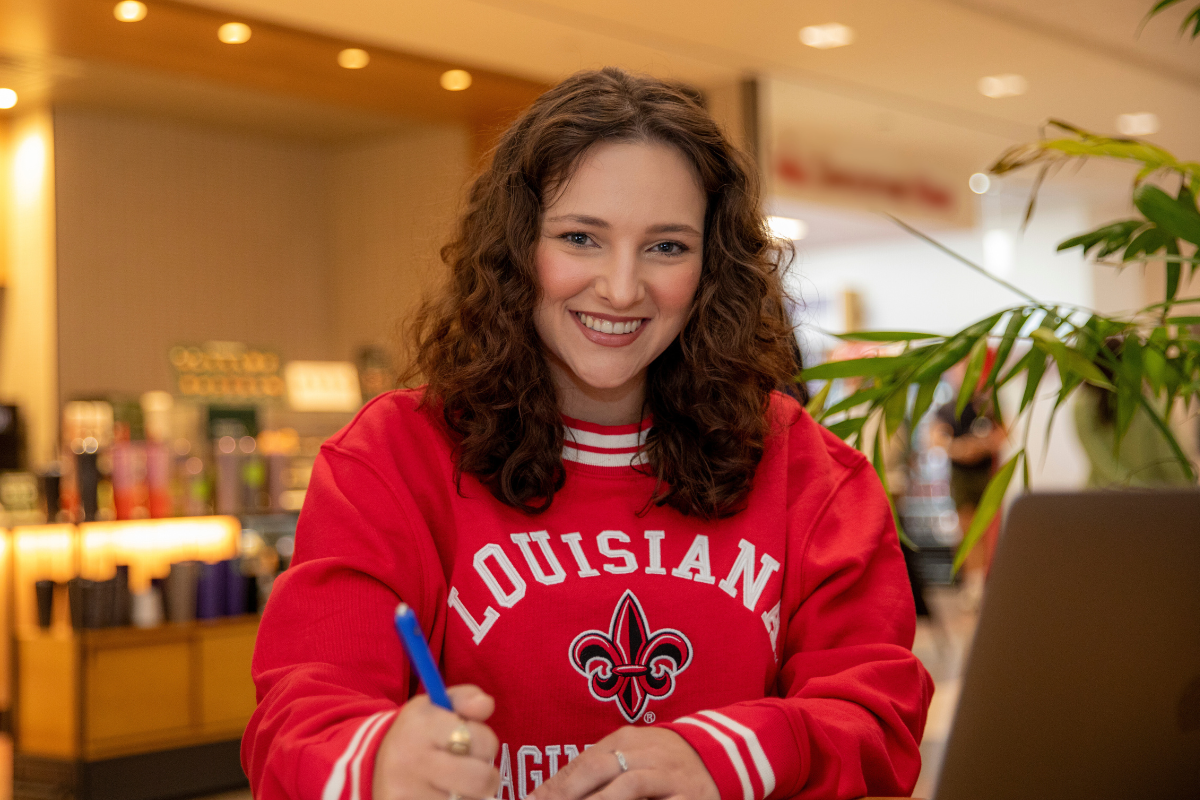 Brooke Perez, a student in the University of Louisiana at Lafayette's online criminal justice degree program, looks up at the camera and smiles as she works on her laptop in a coffee shop. 