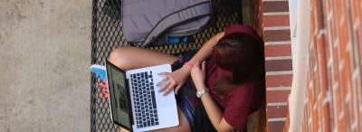 University of Louisiana at Lafayette student sits on campus and looks at her laptop while leaning against a brick wall