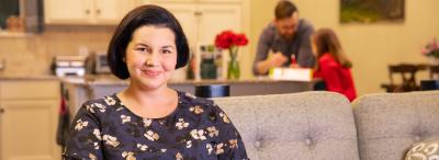 Student sits at laptop in living room with family behind her