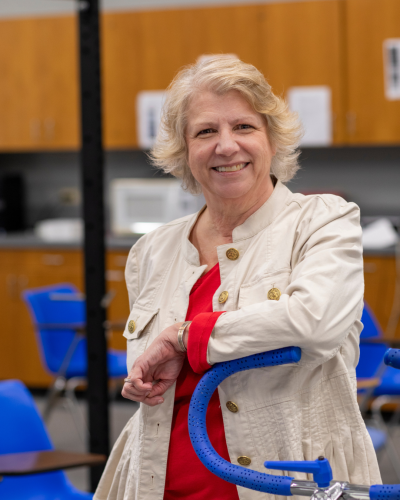 Advisor of UL Lafayette's online bachelor's in kinesiology program smiles, leaning on physical training equipment. She's wearing a red blouse and white coat.