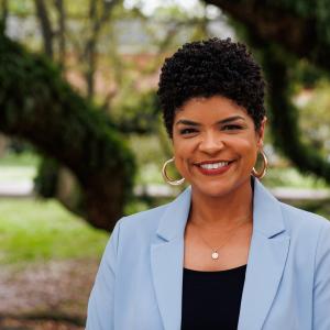 Portrait photo of Marcelle Fontenot in a blue sweater and a black blouse smiling in front of a tree